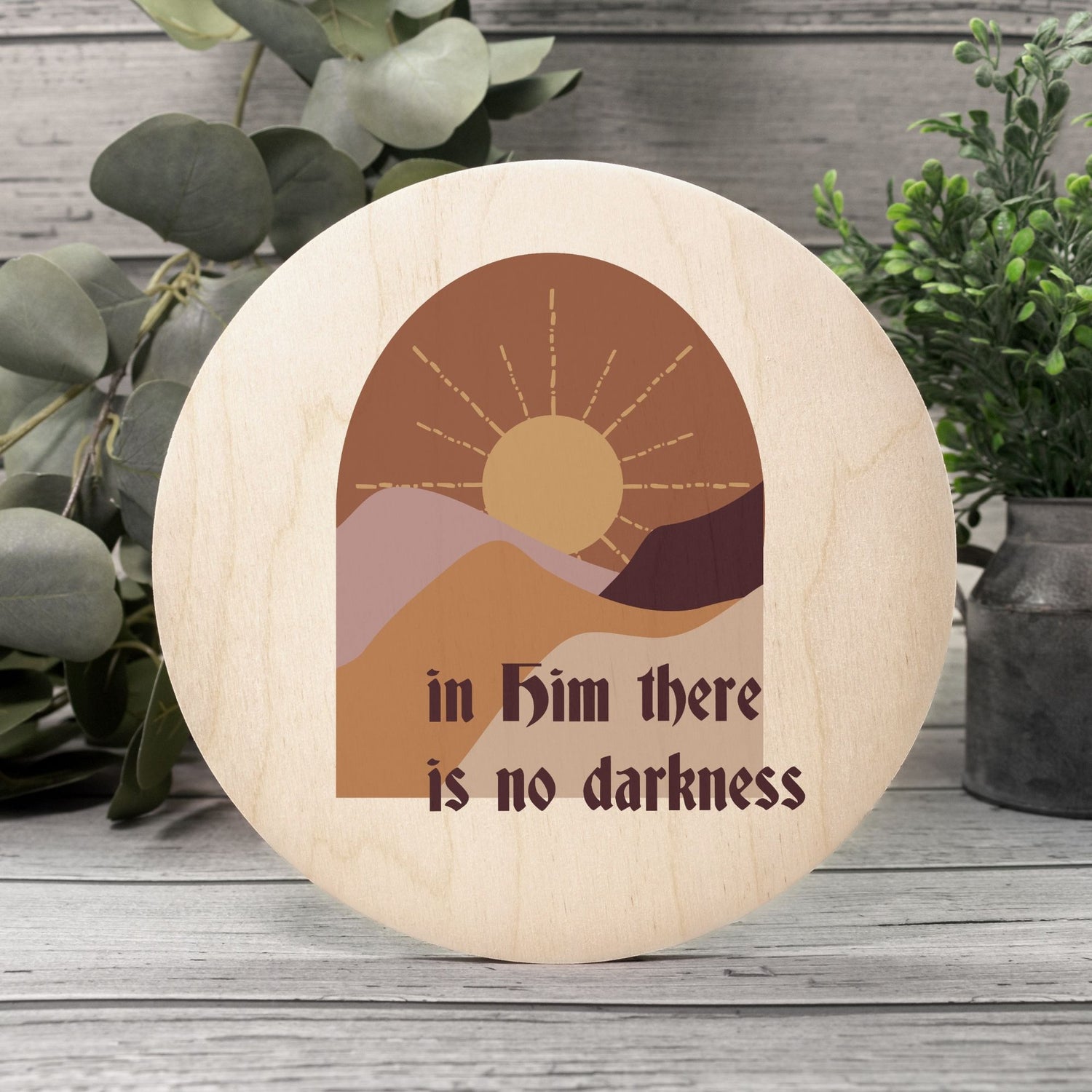 a 10 inch wooden circle piece with a vinyl art print. It is a warm-tones picyre with lots of browns, yellow and orange. A sun rising up over mountains. Minimalistic lines, not realistic. It says "In Him there is no darkness" beneath the mountains.